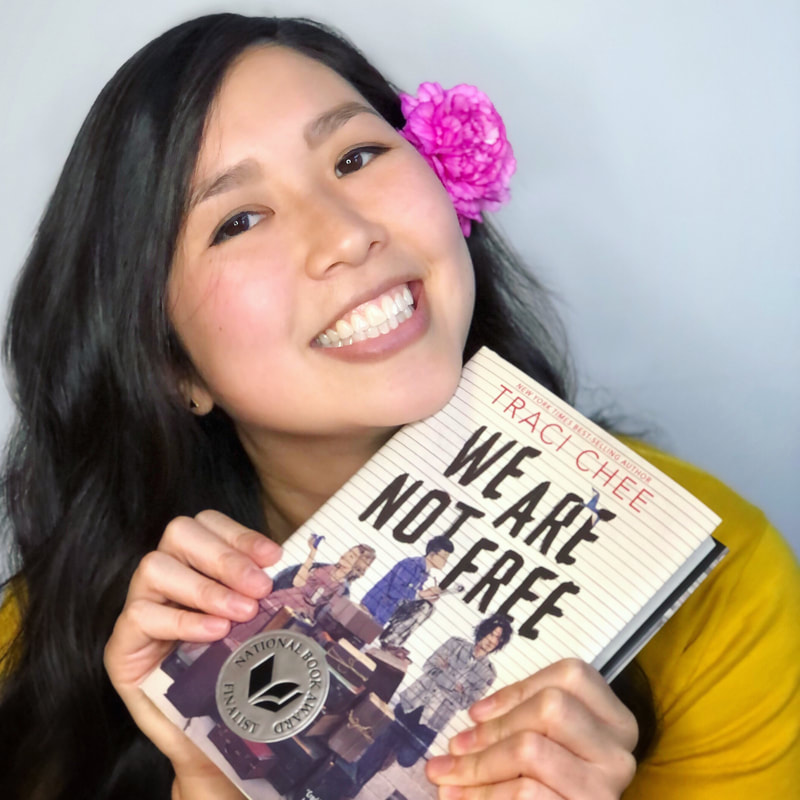Traci Chee in a yellow sweater with a pink flower behind her ear smiling with a copy of We Are Not Free