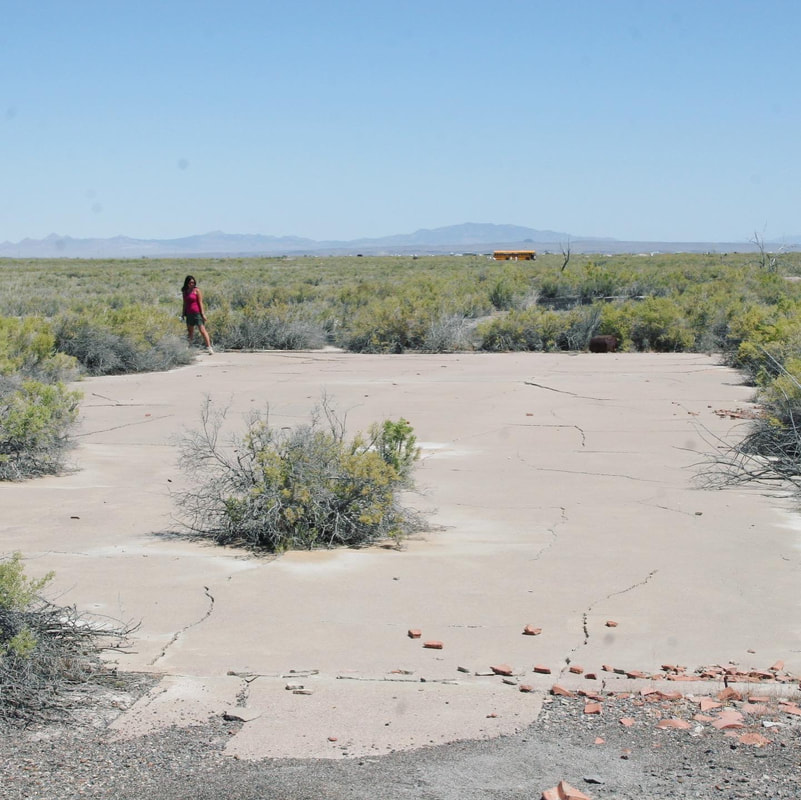 A small female figure in pink standing beside a cracked concrete slab in a scrubby high desert location