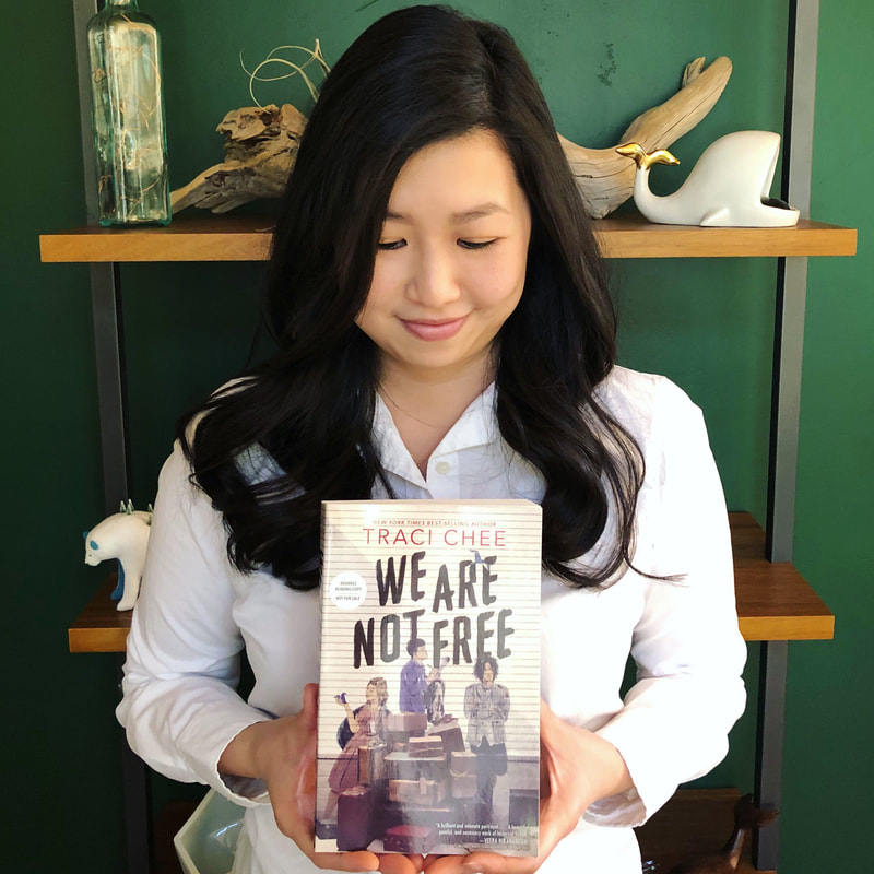 photo of Traci Chee standing in front of a green bookshelf wall with a copy of her book, We Are Not Free