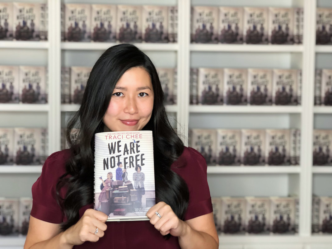Traci Chee in a burgundy blouse holding up a copy of her book We Are Not Free with bookshelves of We Are Not Free behind