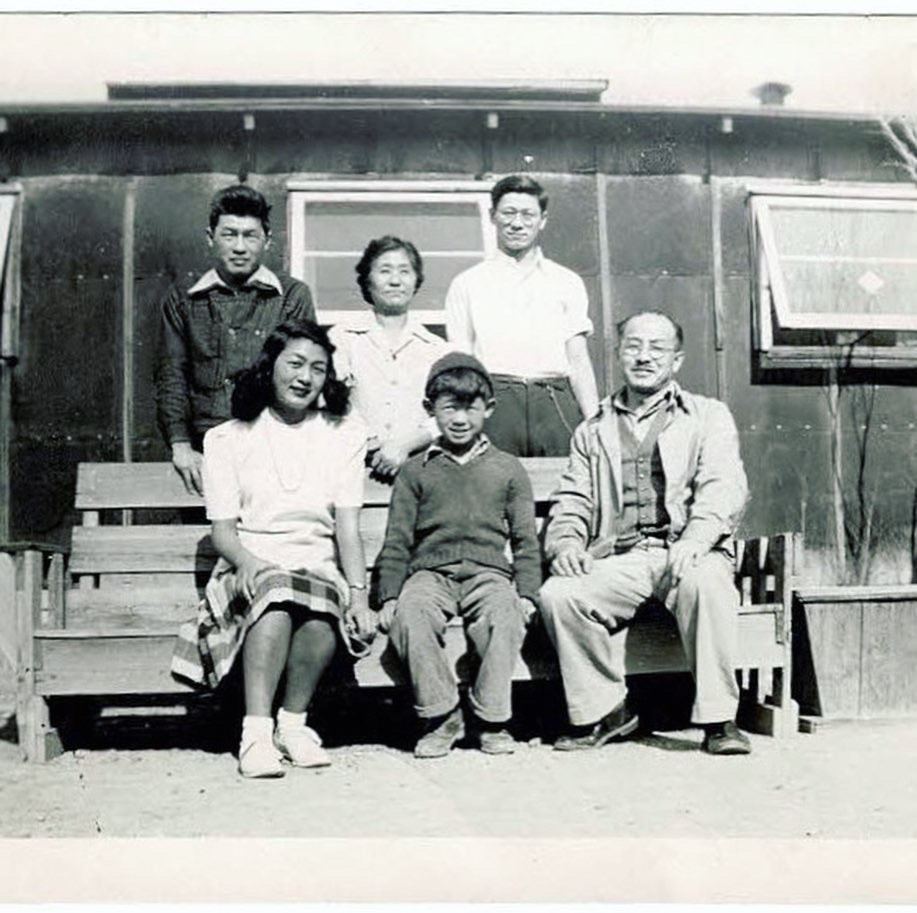 A black and white photo of a Japanese American family sitting on a handmade bench outside of a tar-paper barrack with a potted dead tree behind them