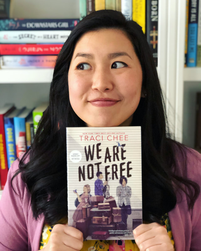 (image: Traci Chee in a purple sweater and yellow floral shirt perching her chin on a copy of her book WE ARE NOT FREE and looking sneaky)