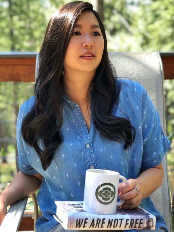 Traci Chee sitting outside in a blue chambray shirt with a copy of We Are Not Free and a white mug with a dark green emblem in her lap