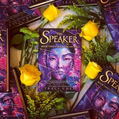 photo of many hardcover editions of THE SPEAKER by Traci Chee with yellow roses and other flowers