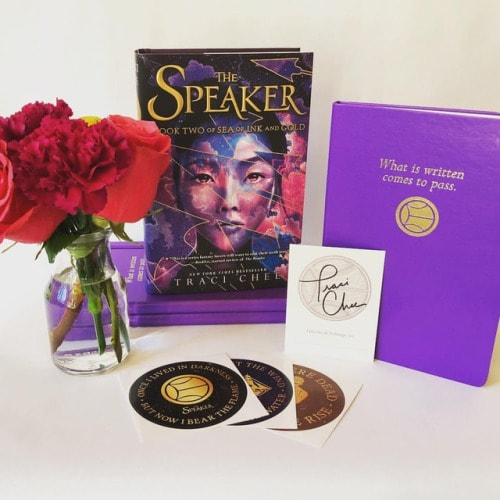 photo of red and pink flowers, THE SPEAKER by Traci Chee, a purple notebook featuring a quote from THE SPEAKER, a signed bookplate, and stickers