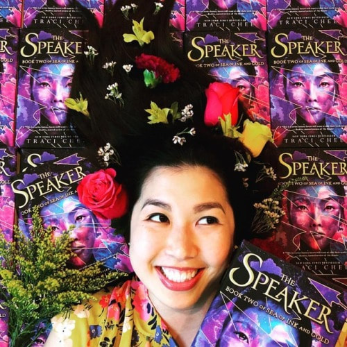 photo of Traci Chee with pink and yellow flowers in her hair, surrounded by copies of THE SPEAKER